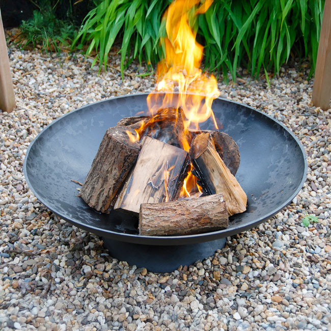 A fire pit in a garden setting with vibrant flames and stacked logs in a metal bowl, surrounded by gravel and lush green plants, creating a cozy atmosphere.
